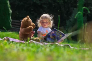 A toddler sitting on a picnic blanket, reading to a stuffed bear.