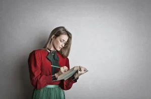 A woman with a burgundy blouse and necktie is wearing glasses and writing in a notebook.