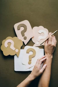 An image of a woman's hands holding a writing pen and 4 paper cut-outs of question marks. 