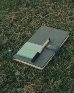 A green notebook on the grass. A pen and a sticky note pad which reads "Here she comes!" are sitting on top of the notebook.