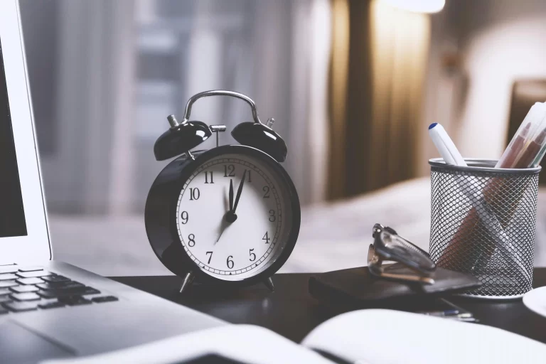 A picture of an alarm clock sitting on a desk next to a laptop and a cup holding several writing pens.