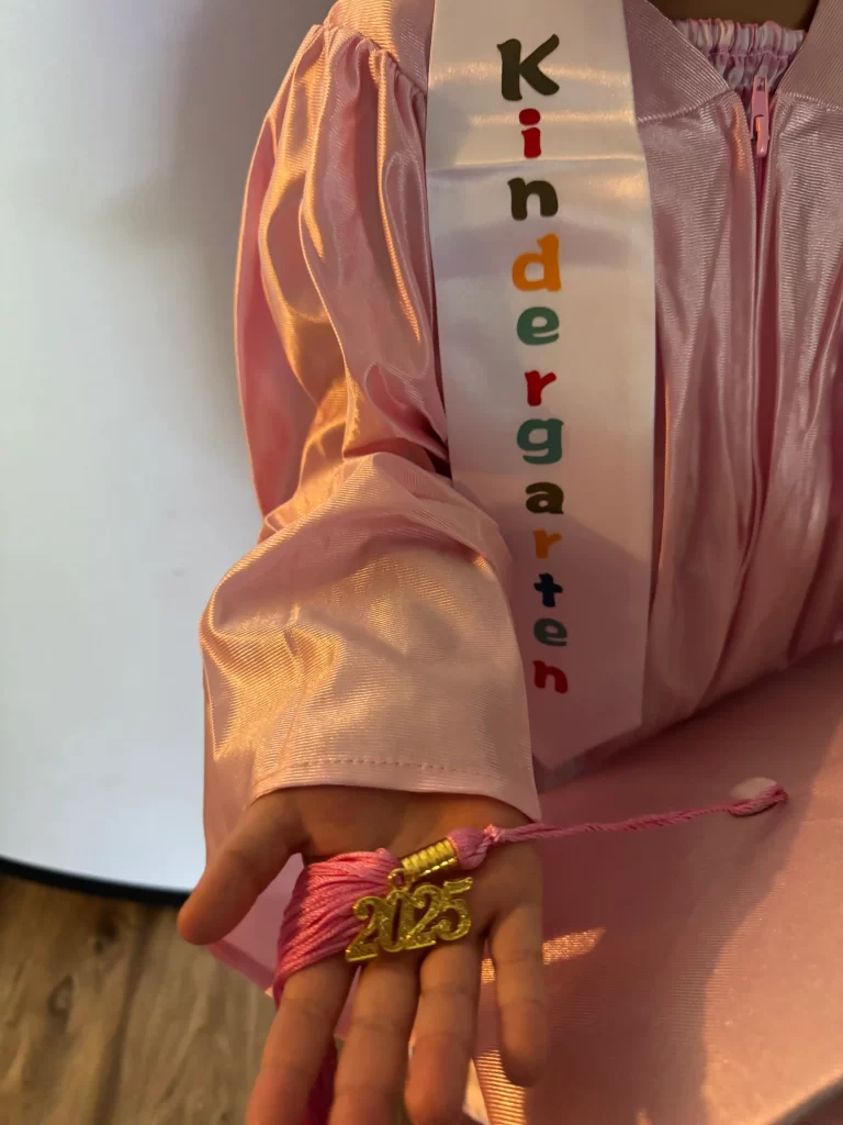 A Kindergarten sash laying over a child's shoulder. The child is wearing a pink gown and holding the 2025 tassle in her hand.