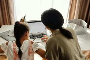 A mom working on a laptop while her daughter sits next to her, drawing a picture.