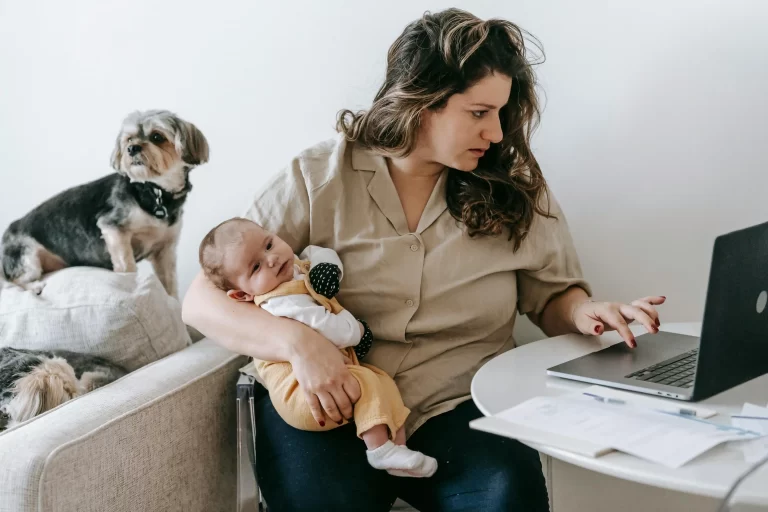A mother working on a computer, holding a baby with a dog in the background.