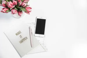 A cellphone, pen and notebook on a white table. A vase with pink roses sits on the table too. The words on the notebook read "make it happen".