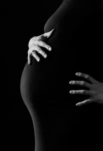 A pregnant person, wearing a black dress, holding their stomach against a black background.