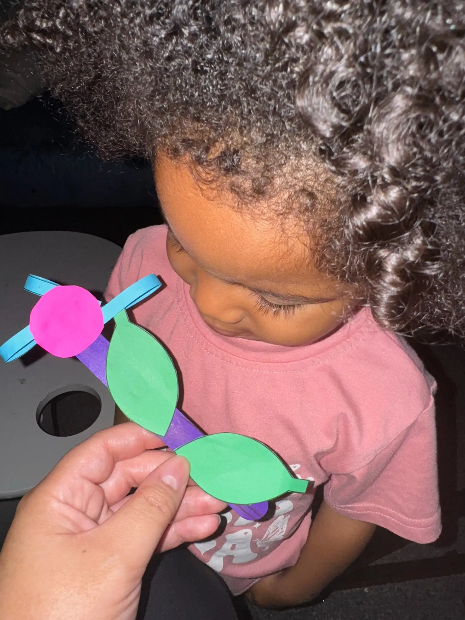 A toddler girl looking at a flower made of a popsicle stick and construction paper.