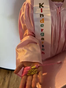 A Kindergarten sash laying over a child's shoulder. The child is wearing a pink gown and holding the 2025 tassle in her hand.