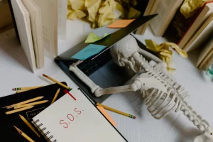 A skeleton leaned over a laptop. On his notebook is written 'SOS', representing purposeful morning routine homeschoolers.
