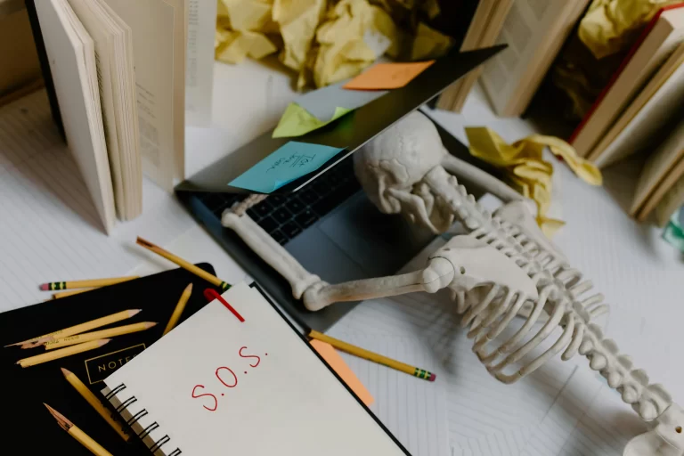 A skeleton leaned over a laptop. On his notebook is written 'SOS', representing purposeful morning routine homeschoolers.